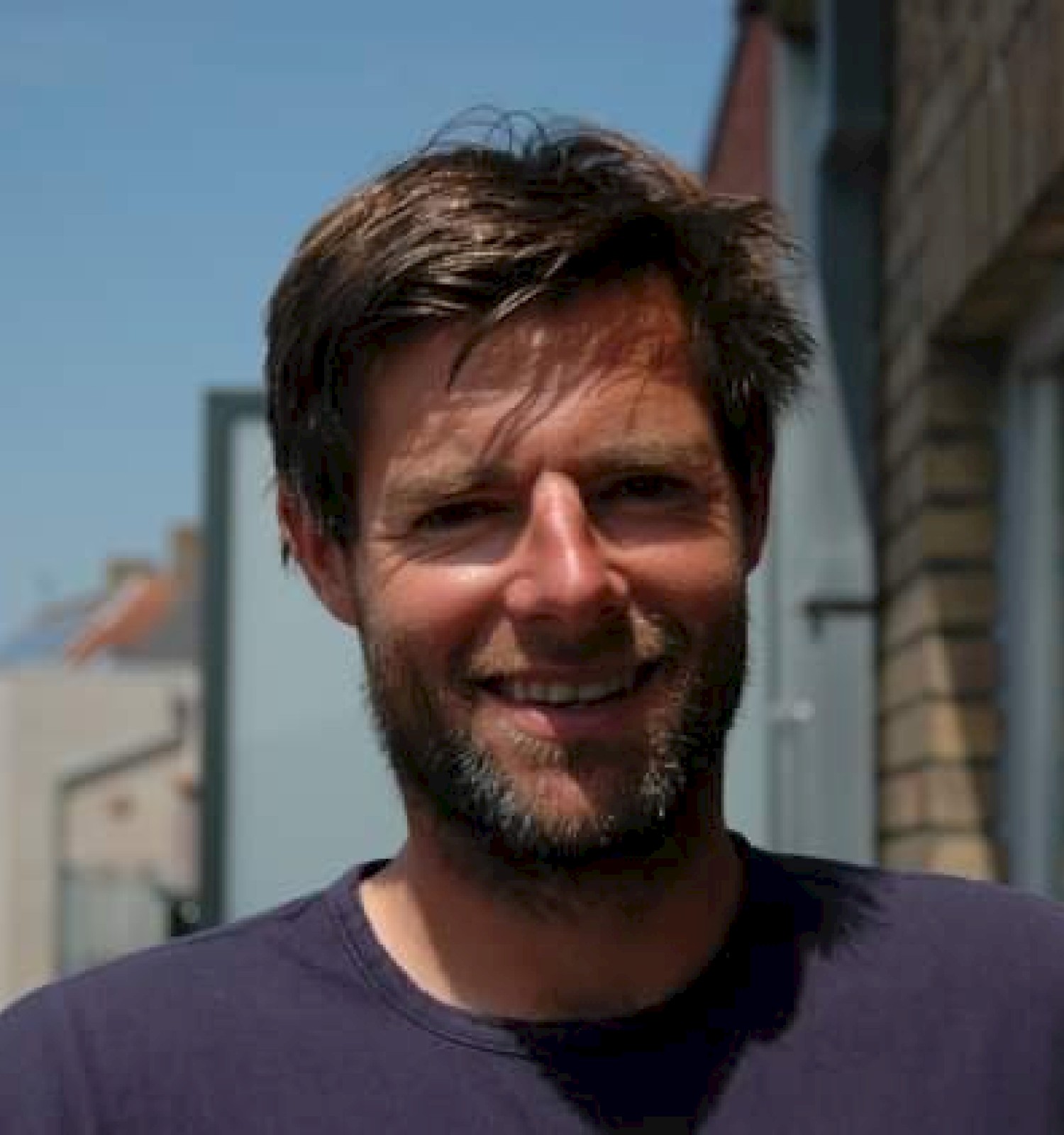A man with brown hair and a beard smiles at the camera outside, standing near a brick building on a sunny day.