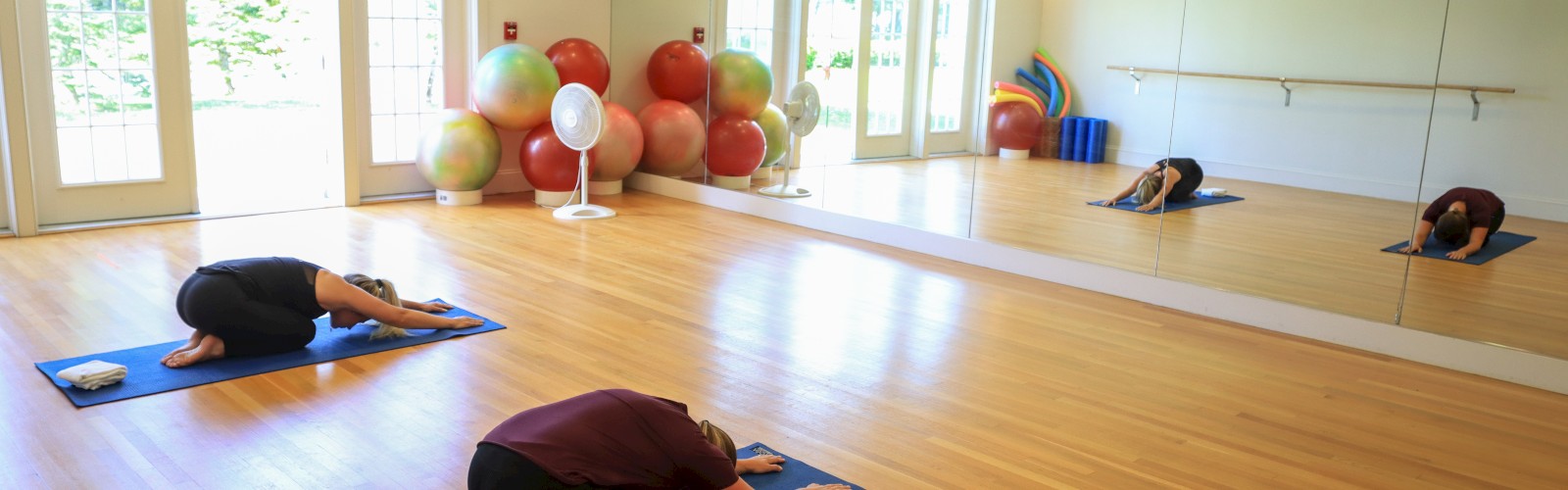 Three people are practicing yoga in a room with wooden floors, large mirrors, yoga mats, and exercise balls in the background, by the door.