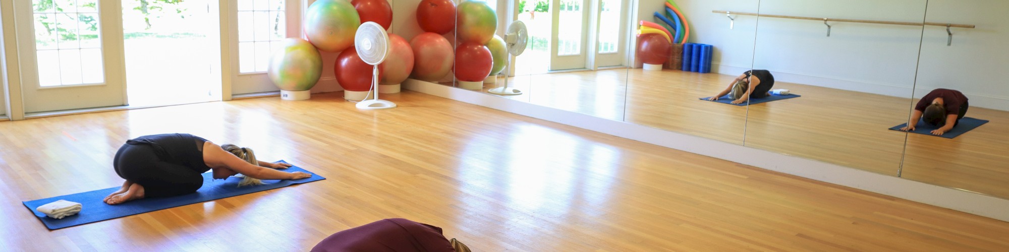 Three people are practicing yoga in a room with wooden floors, large mirrors, yoga mats, and exercise balls in the background, by the door.