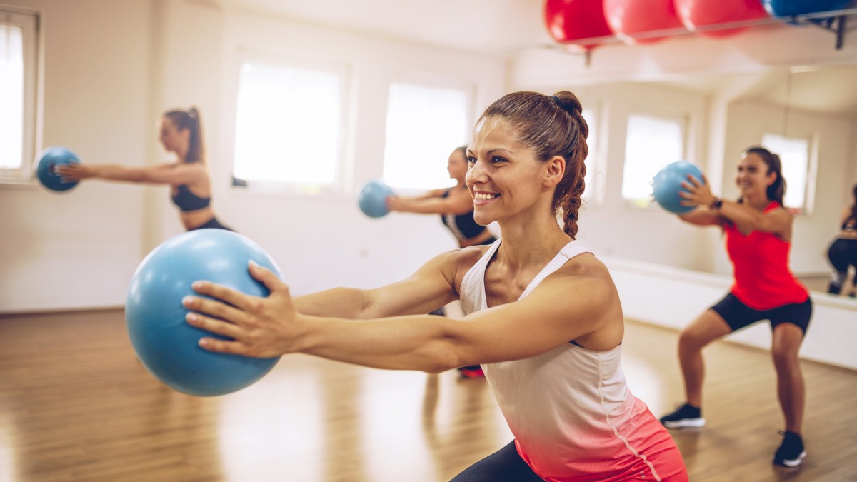 A group of women are participating in a fitness class, holding blue exercise balls and performing squats in a mirror-walled studio with wooden floors.