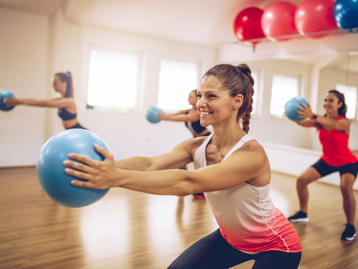 A group of women are participating in a fitness class, holding blue exercise balls and performing squats in a mirror-walled studio with wooden floors.