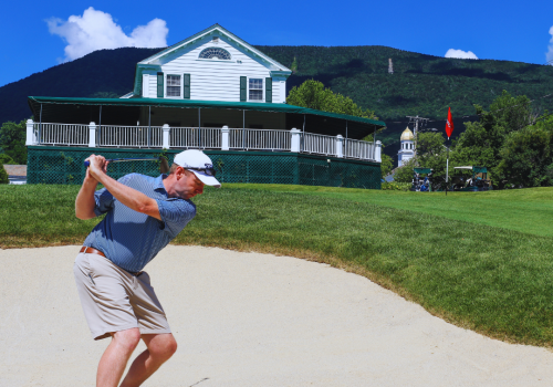 A man is playing golf, preparing to hit the ball from a sand trap with a white building and mountains in the background, under a clear blue sky.