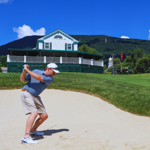 A man is playing golf, preparing to hit the ball from a sand trap with a white building and mountains in the background, under a clear blue sky.