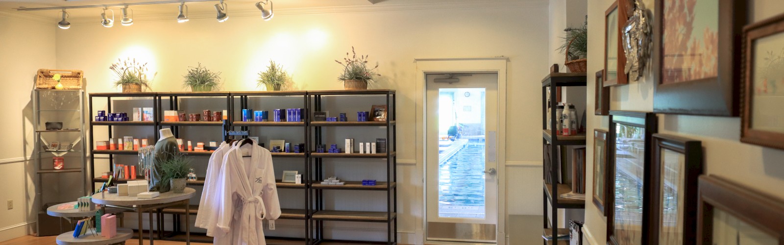 The image shows a well-lit wellness center interior with wooden flooring, shelves with products, robes on display, and framed pictures on the wall.