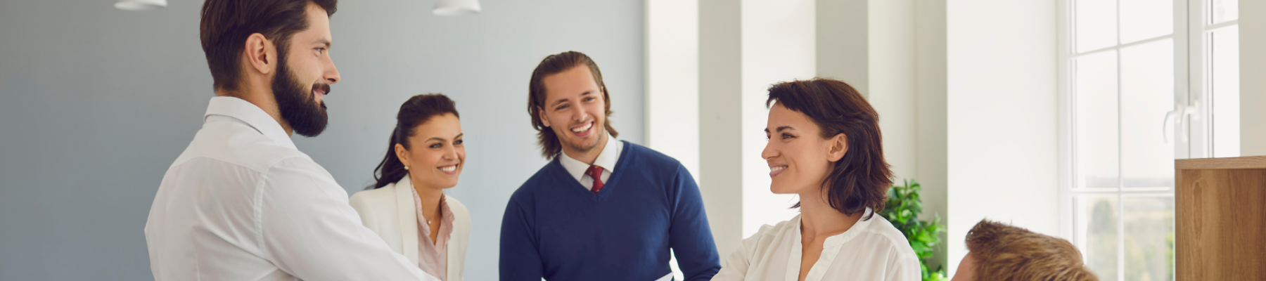 A business meeting scene where two people are shaking hands, while three others observe and smile, in a bright, modern office setting.