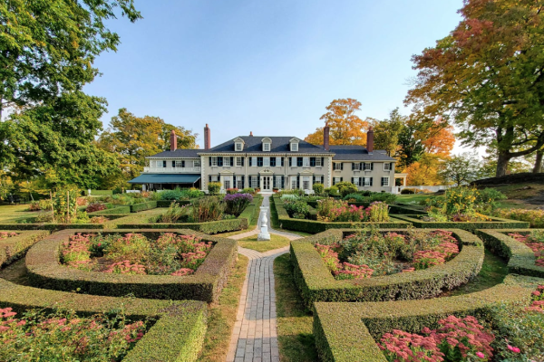 A large, elegant estate with a beautifully manicured garden featuring intricate hedges, a central pathway, and surrounded by colorful flowers and trees.