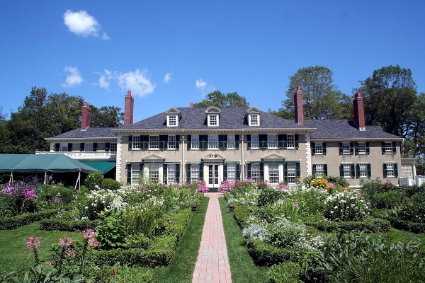 The image shows a large, elegant mansion with multiple chimneys, surrounded by beautifully landscaped gardens and a brick walkway leading to the entrance.