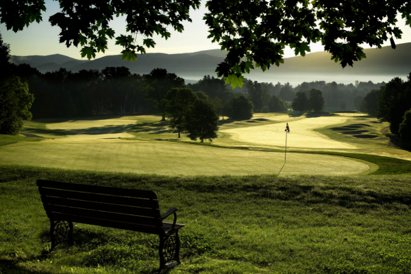 A scenic view of a golf course with a bench in the foreground, surrounded by trees and mountains under a clear sky at dawn or dusk.