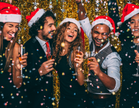 A group of five people wearing Santa hats are celebrating, holding drinks and smiling with a gold sequin background and festive atmosphere.