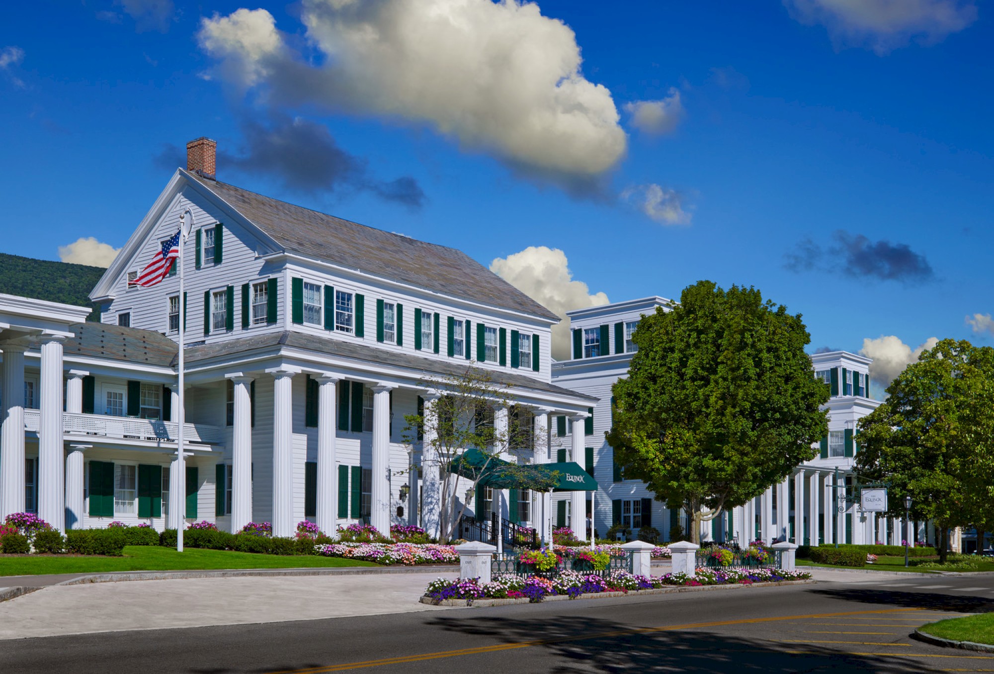 A large, white colonial-style building surrounded by flowers and trees, with an American flag flying in front, under a blue sky with scattered clouds.