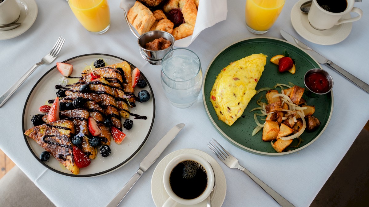 A breakfast spread with pancakes topped with berries, an omelet, potatoes, croissants, muffins, coffee, orange juice, and water.