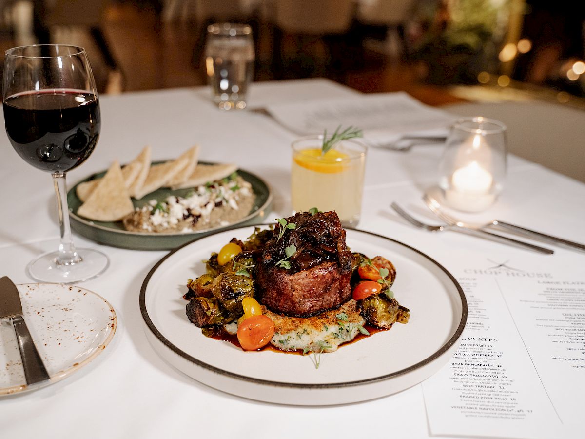 A gourmet meal on a table with beef, vegetables, red wine, a cocktail, soft tacos, and a menu in view next to a lit candle, ready to be enjoyed.