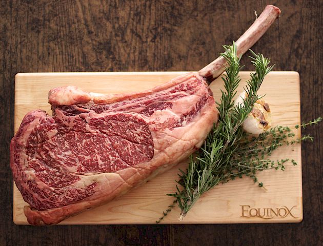 A large, marbled cut of raw beef with a bone, garnished with fresh herbs, placed on a wooden cutting board marked 