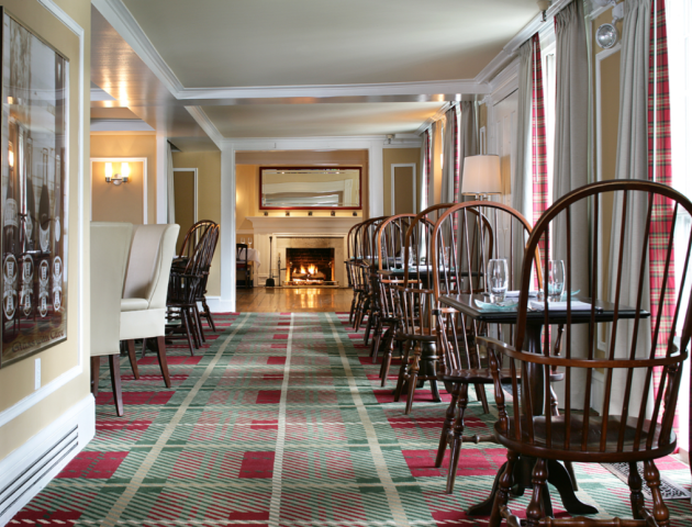 This image shows a cozy restaurant interior with wooden chairs and tables, a patterned carpet, large windows, and a lit fireplace in the background.