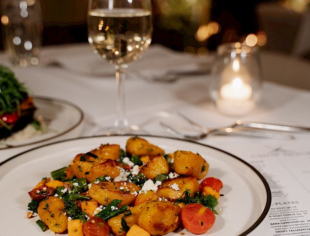 The image shows a plate of roasted vegetables on a table, accompanied by a glass of white wine, with a candle and a menu beside it.