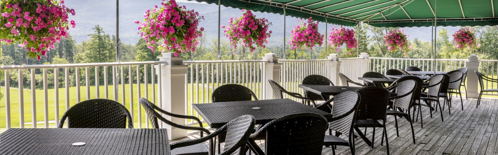 Outdoor patio with black tables and chairs under a green canopy, pink flowers hanging from the railing, and a scenic view.