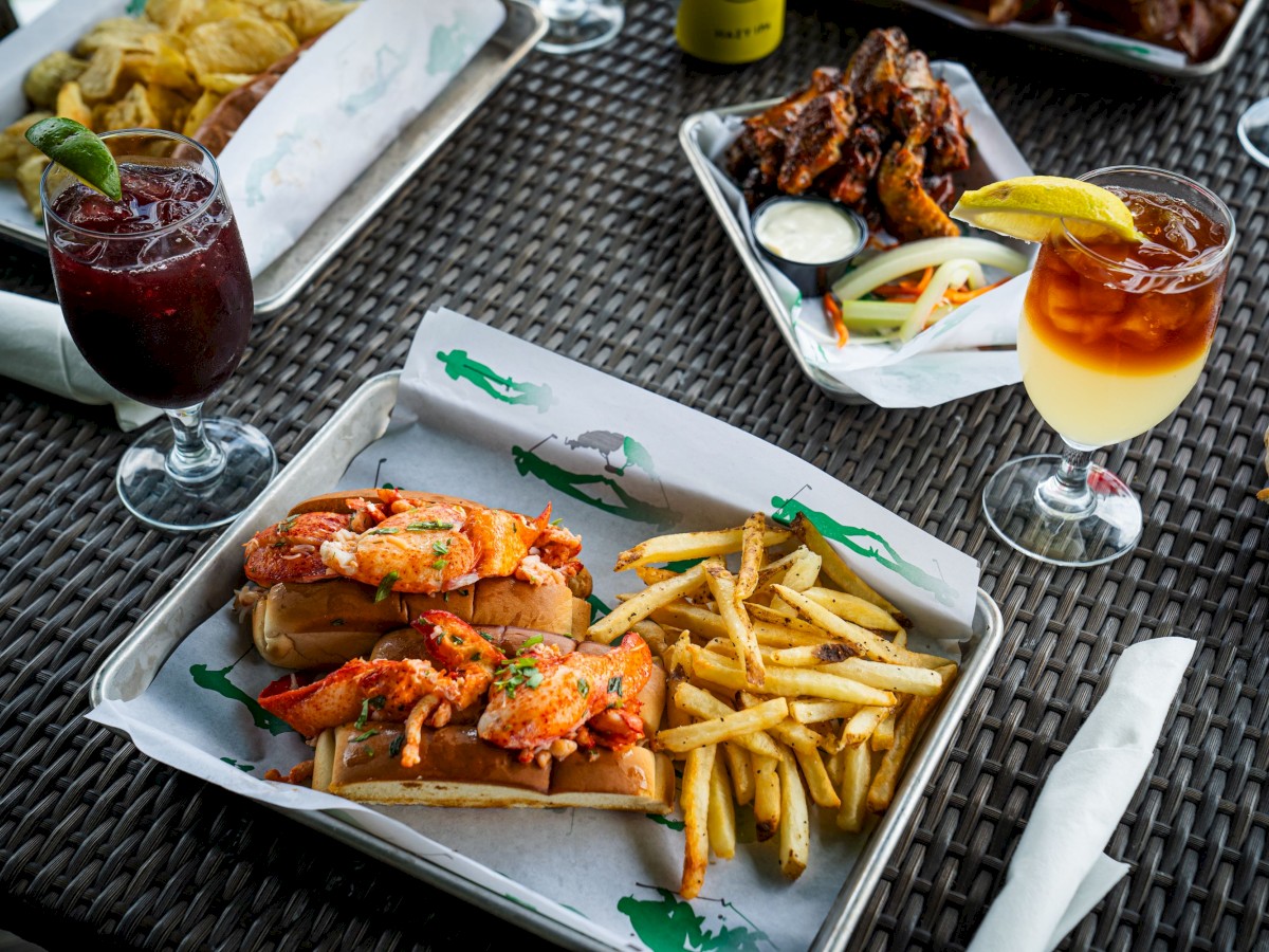 The image shows a table with lobster rolls and fries, chicken wings, drinks with garnishes, and a plate of chips. Everything is served on trays.