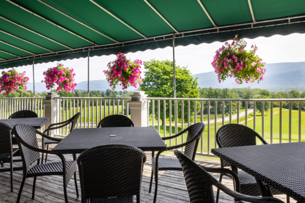 The image shows a patio with black wicker furniture under a green canopy. Colorful hanging flower baskets are along the railing, and a scenic background is visible.