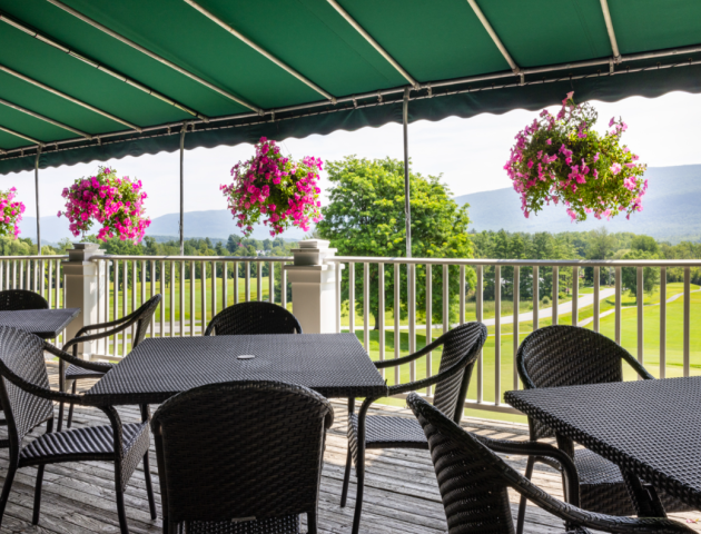 The image shows a patio with black wicker furniture under a green canopy. Colorful hanging flower baskets are along the railing, and a scenic background is visible.