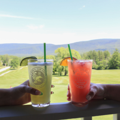 Two hands holding drinks with straws and garnishes on a balcony, overlooking a scenic view of trees, hills, and a golf course.