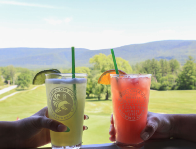 Two hands holding drinks with straws and garnishes on a balcony, overlooking a scenic view of trees, hills, and a golf course.