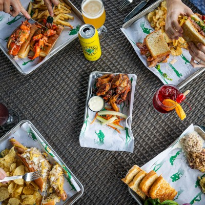 Five people are enjoying various meals and drinks around a table, featuring sandwiches, chips, wings, and beverages, all on a woven surface.