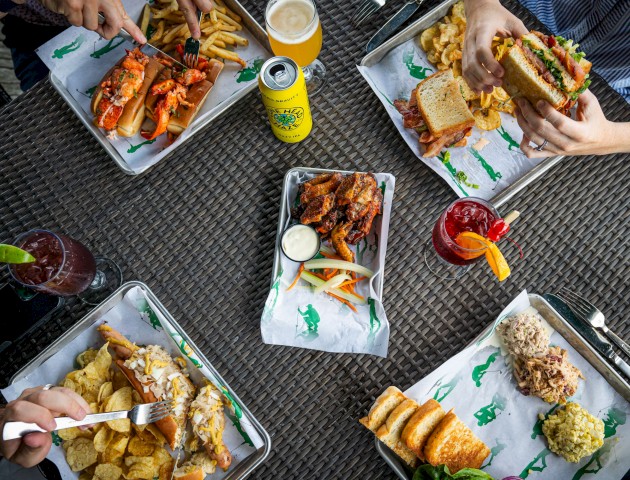 Five people are enjoying various meals and drinks around a table, featuring sandwiches, chips, wings, and beverages, all on a woven surface.