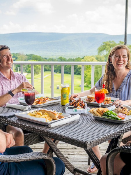 Four people enjoying a meal on a deck with scenic mountain views, smiling, and having drinks around a table.
