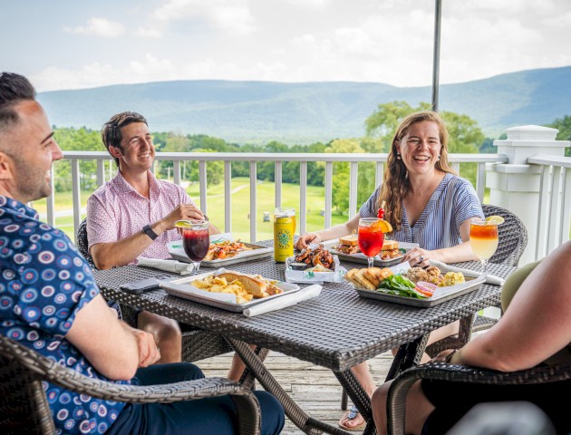 Four people enjoying a meal on a deck with scenic mountain views, smiling, and having drinks around a table.