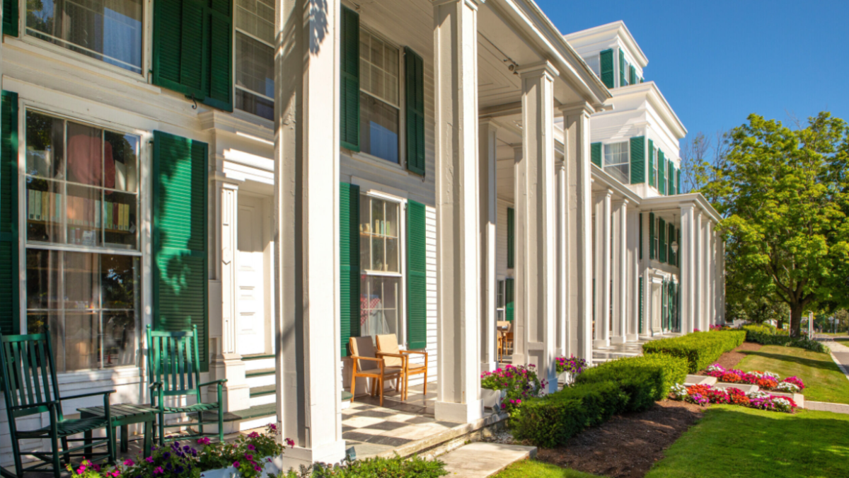 A row of white-columned buildings with green shutters and rocking chairs, surrounded by a well-maintained garden and walkway, under a clear blue sky.
