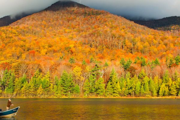 Two people are fishing in a small boat on a lake during autumn, with a colorful forest and mountain in the background.