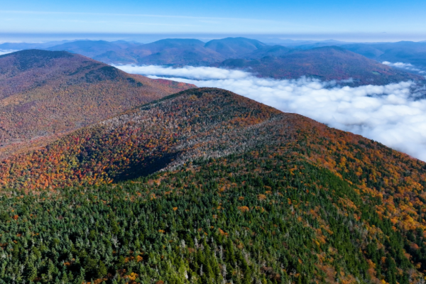 An aerial view of mountains with autumn foliage, showcasing green and orange trees, under a blue sky with scattered clouds in the background.