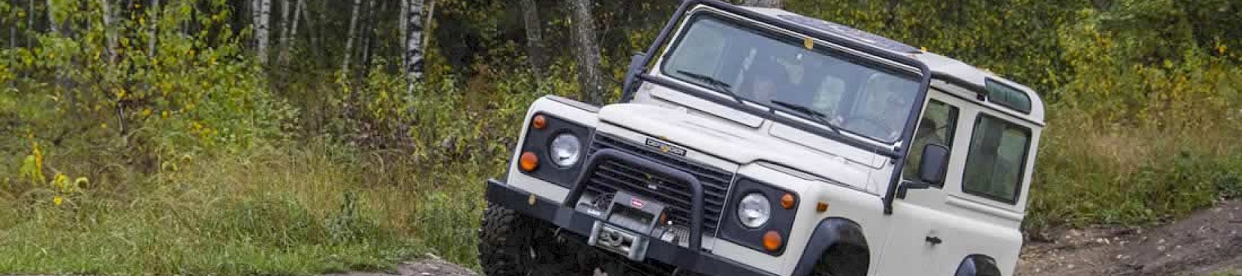 A white off-road vehicle is navigating a muddy path in a forested area with trees displaying autumn colors.