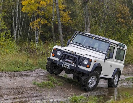 A white off-road vehicle is navigating a muddy path in a forested area with trees displaying autumn colors.