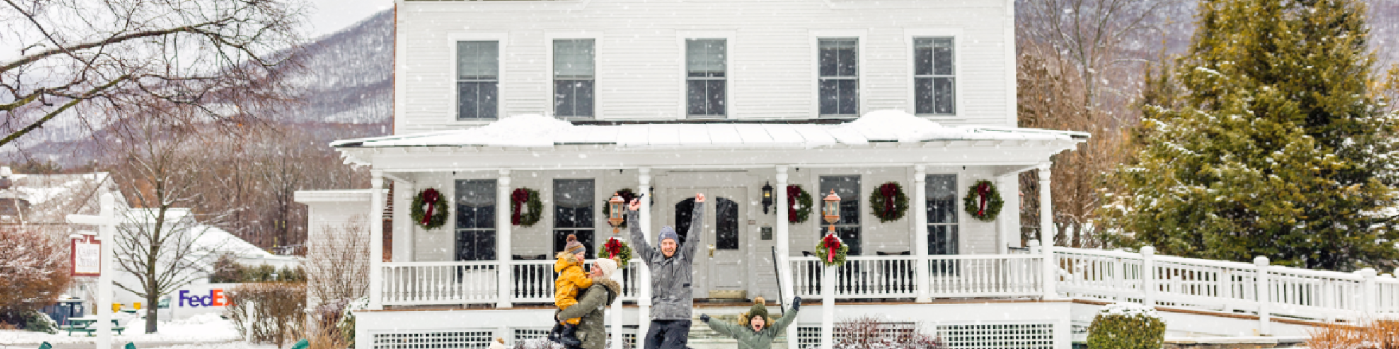 A family is playing in the snow in front of a large, white house adorned with Christmas wreaths, while other houses and trees are visible in the background.