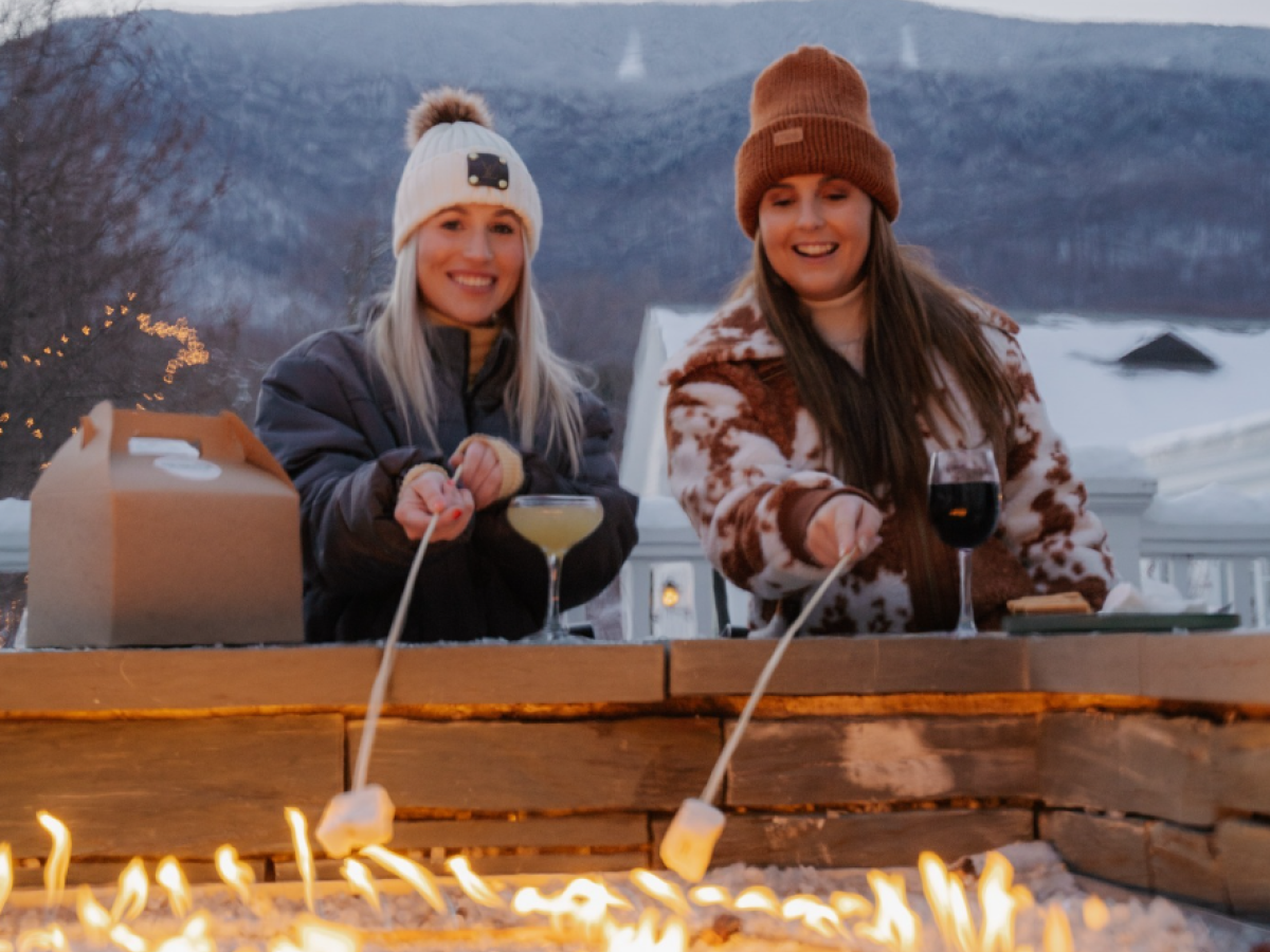 Two people are roasting marshmallows over an outdoor fire pit with snowy mountains in the background, holding drinks and smiling.