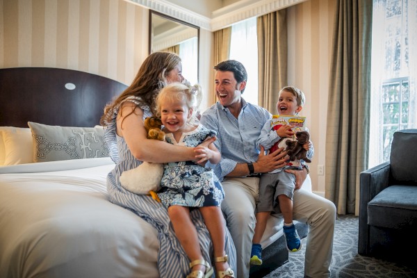 A family of four is sitting on a bed in a well-lit room, smiling and holding stuffed animals and a book.