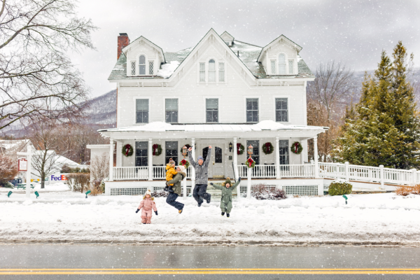 A family jumps in the snowy street in front of a large, decorated white house with wreaths, providing a festive winter scene.