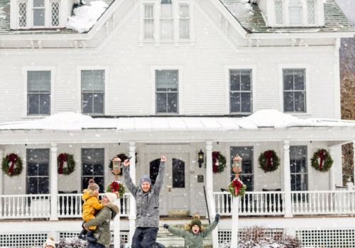 A family of four joyfully plays in the snow outside a large white house decorated with wreaths, capturing a festive winter scene.