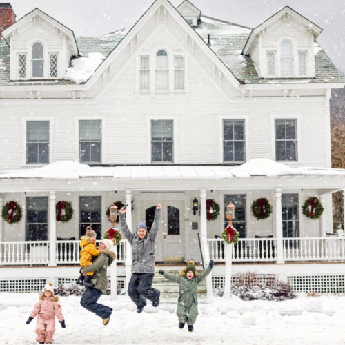 A family of four joyfully plays in the snow outside a large white house decorated with wreaths, capturing a festive winter scene.