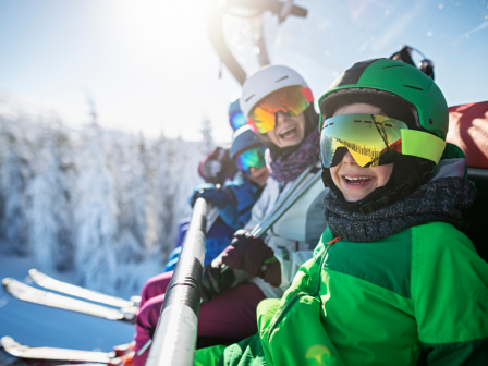 A group of people on a ski lift, wearing helmets and goggles, surrounded by snow-covered trees and a bright, sunny sky.