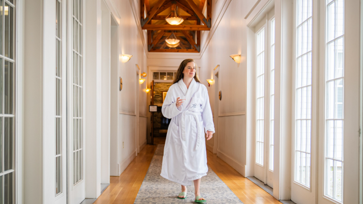 A person in a white robe and slippers walks down a bright hallway with wooden floors and large windows.