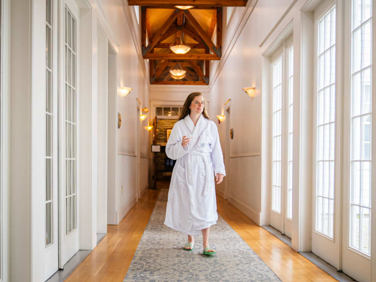 A person in a white robe and slippers walks down a bright hallway with wooden floors and large windows.