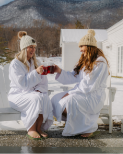 Two people in winter hats and robes sit outdoors, clinking mugs. Snow covers the ground, with mountains in the background.