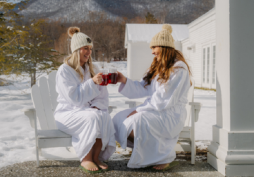 Two people in winter hats and robes sit outdoors, clinking mugs. Snow covers the ground, with mountains in the background.