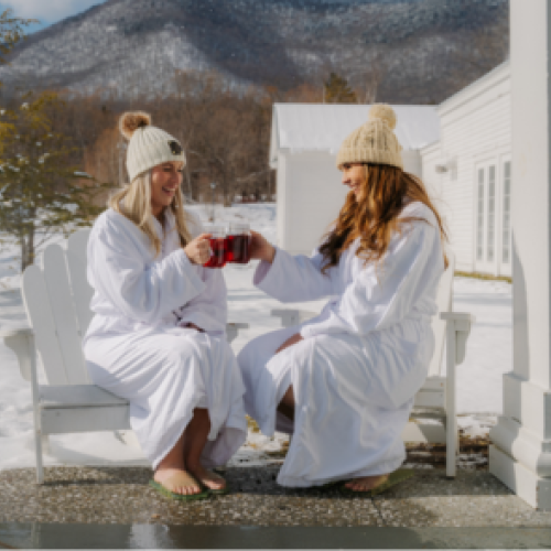 Two people in winter hats and robes sit outdoors, clinking mugs. Snow covers the ground, with mountains in the background.