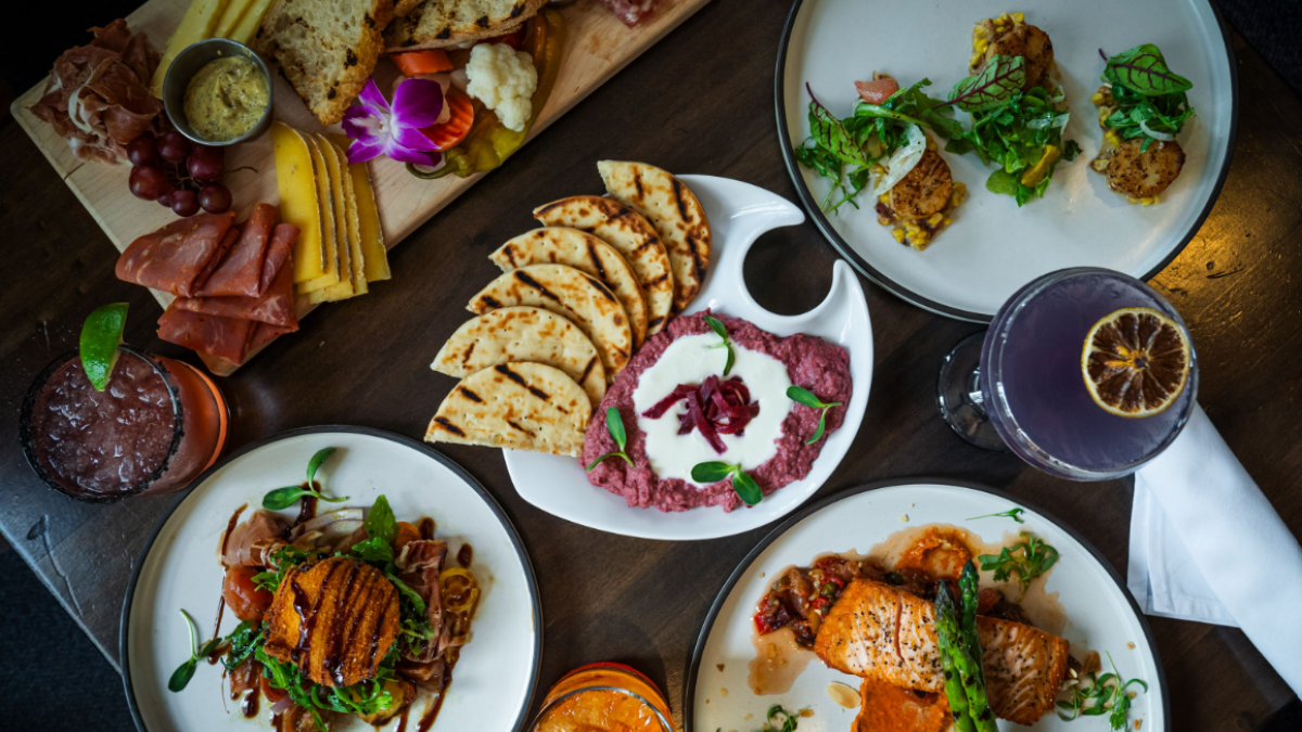 The image shows a variety of dishes including meats, cheeses, pita with dip, and drinks on a wooden table.