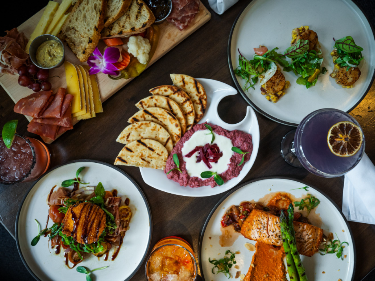 The image shows a variety of dishes including meats, cheeses, pita with dip, and drinks on a wooden table.