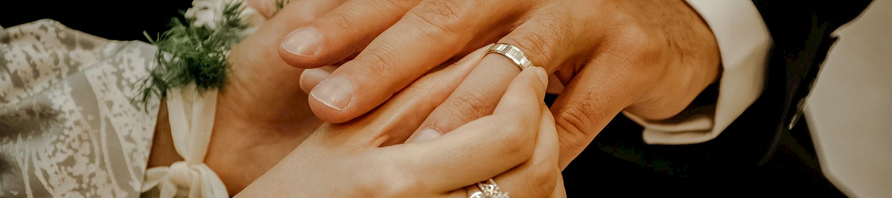 Two hands with rings are shown in a close-up, possibly during a wedding ceremony, with a floral detail visible on one wrist.