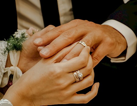 Two hands with rings are shown in a close-up, possibly during a wedding ceremony, with a floral detail visible on one wrist.